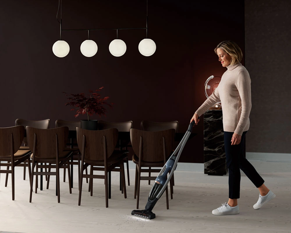 A person vacuuming the floor with an Electrolux cordless stick vacuum in a modern dining room featuring dark walls, wooden dining chairs, and contemporary pendant lighting.