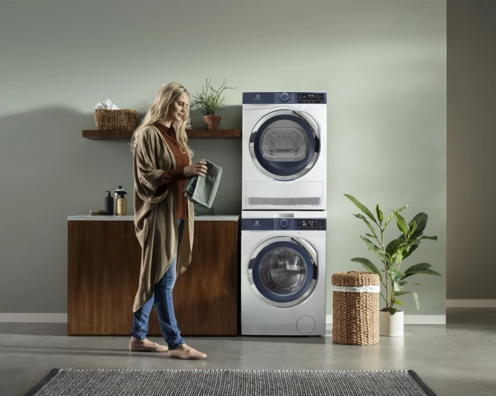 A modern laundry space with a person standing in front of a stacked Electrolux washer and dryer, next to a wooden cabinet with neatly arranged baskets, plants, and laundry items.